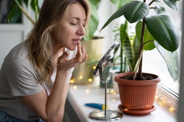 Woman applying moisturizing nourishing balm to her lips with her finger to prevent dryness and chapping in the cold season, sitting by the window, looking at mirror. Lip protection.