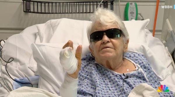 A white white-haired woman with cataract glasses on lies in a bed with white gauze on her hand.