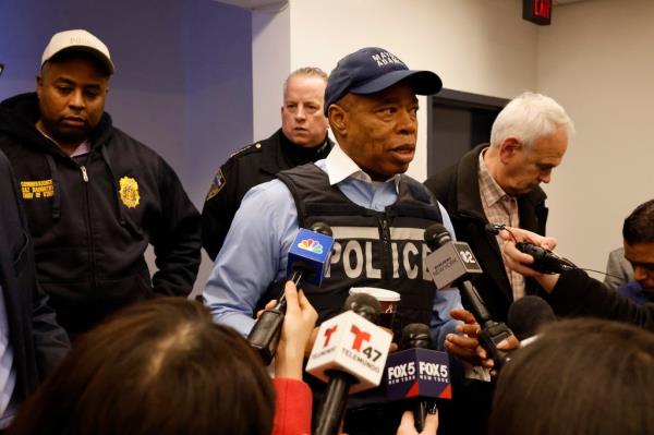 Mayor Eric Adams speaking into micropho<em></em>nes while wearing a blue cap and vest at the 28th precinct in NYC, fighting against the 