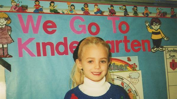 JonBenét Ramsey seen smiling in front of classroom kindergarten banner.