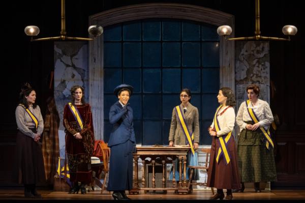 Nikki M. James portraying Ida B. Wells in a scene from the Broadway show 'Suffs', surrounded by other actresses on stage.