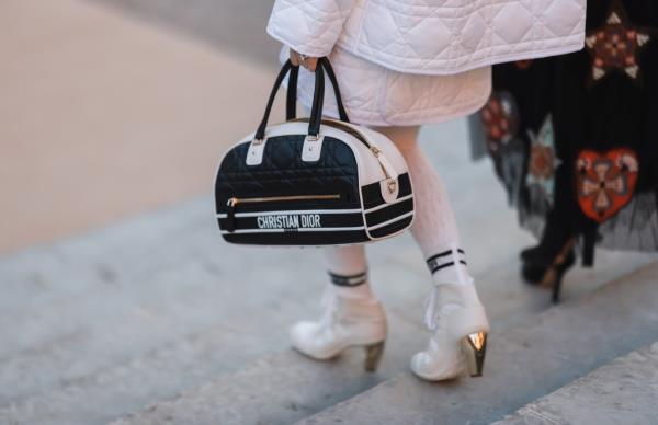 A guest at Paris Fashion Week wearing a white quilted Dior jacket, a matching short skirt, white cut-out tights, and holding a black and white Dior Vibe leather handbag