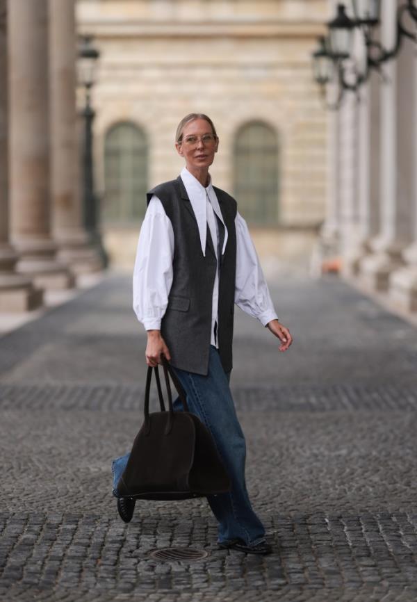 Mirja Klein in Munich, wearing aviolet glasses, oversized grey waistcoat, white shirt, wide-legged jeans, with a large brown suede bag and black leather loafers