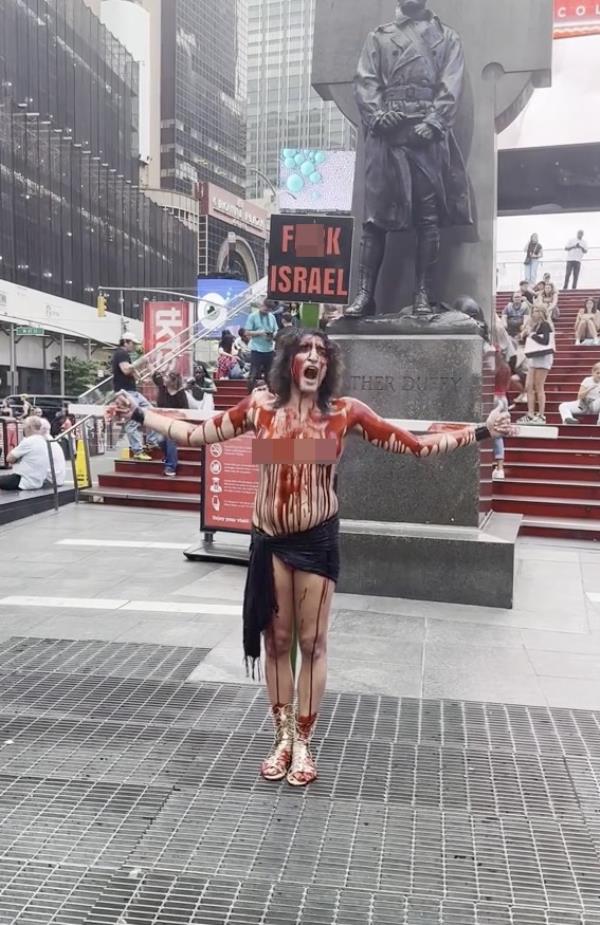 A topless woman drenched in fake blood strapped to a crucifix holding a sign that says f--k israel.