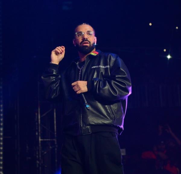 Drake stands on a stage in a leather jacket and clear glasses with a blank ex<em></em>pression on his face