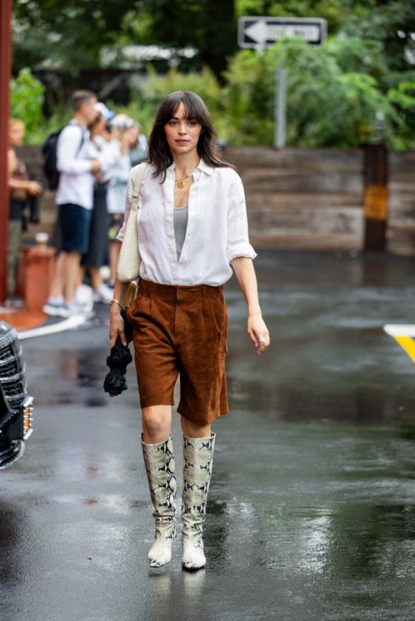 A woman wearing brown leather shorts, snake print boots, and a white button shirt walking on a wet street during New York Fashion Week in September 2023