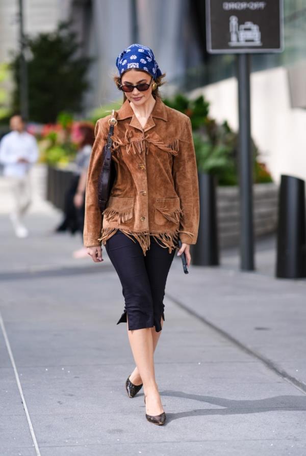 A guest at New York Fashion Week 2024 wearing a dark blue patterned head scarf, gold earrings, a fringe suede jacket, a black leather bag with gold handle, black capri pants and white pointed-toe heels