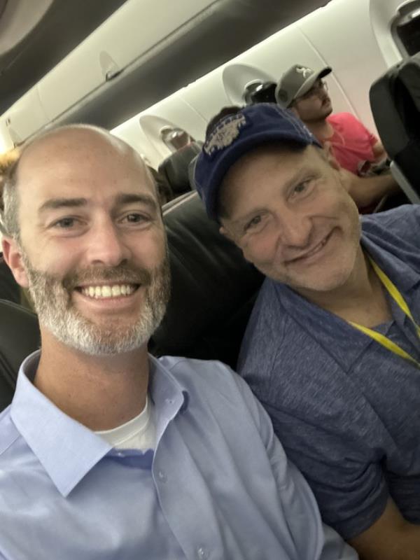 Photo of two men smiling together on a plane. 