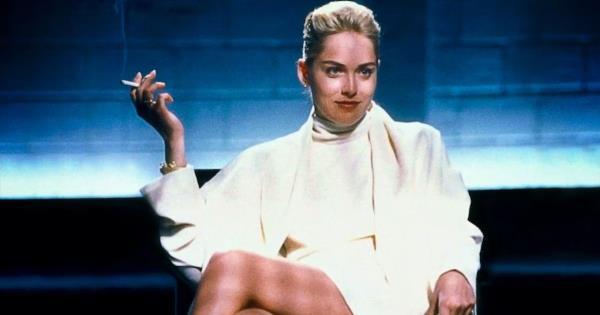 Sharon Stone wearing a white coat in a blue room with legs crossed, smoking.