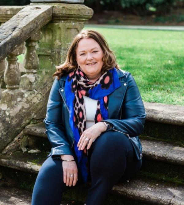 Alison Rios McCrone sitting on stone steps outside, with a stone balustrade to her right and grass in the background.