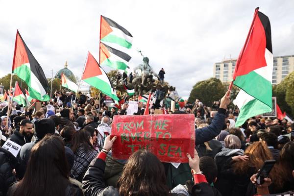 Protester in Berlin raising a placard that reads 'From the river to the sea' during a solidarity rally with Palestinians, while people around hold signs and flags.