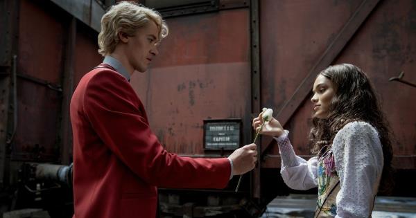 Tom Blyth as Coriolanus Snow in a red suit holds out a flower to a young woman, Rachel Zegler, as Lucy Gray Baird..