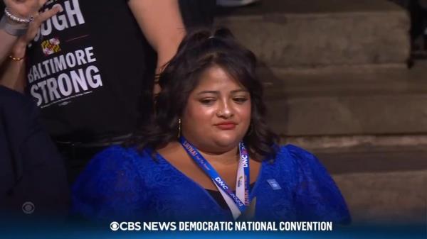 Camera pans over to a woman in a blue dress appearing to be standing somewhat uncomfortably in the crowd.