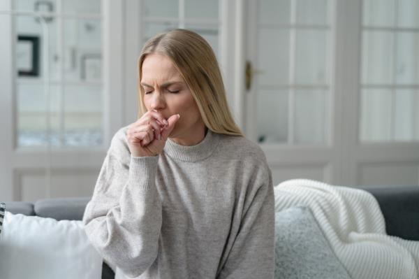 upset blo<em></em>nde woman with fever and cough holding hand to her mouth, feeling weak and alone at home with bro<em></em>nchitis and viral infection.