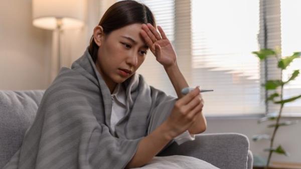 A sick young woman wrapped in a blanket on her couch, taking her temperature